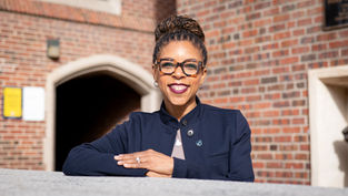 Smiling woman in blue jacket with glasses leans on a gray surface. Brick wall and archway in the background. Bright, sunny day.