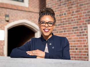 Smiling woman in blue jacket with glasses leans on a gray surface. Brick wall and archway in the background. Bright, sunny day.