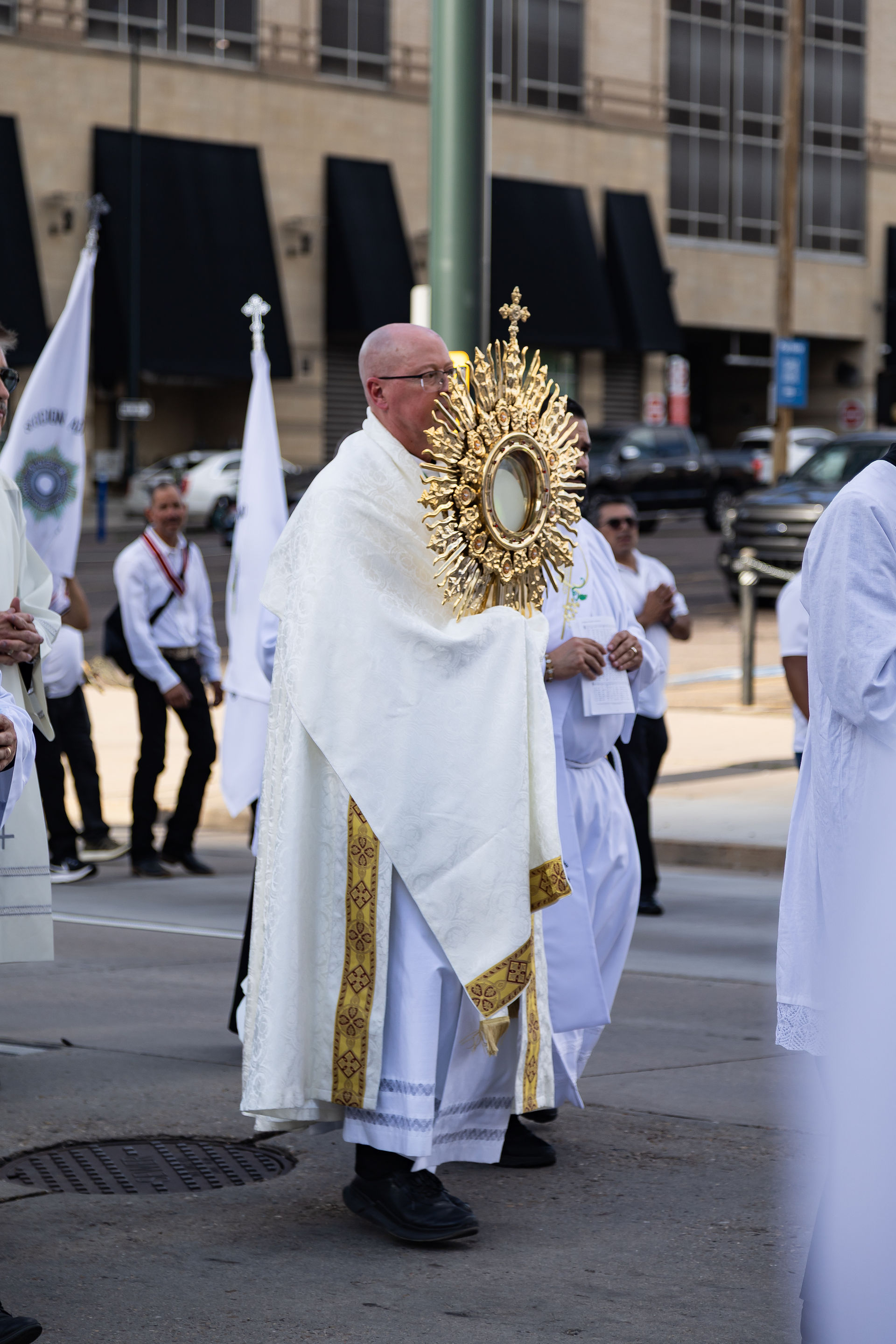 Priest in white robes carries a golden monstrance during a solemn street procession, with other clergy and flags behind.