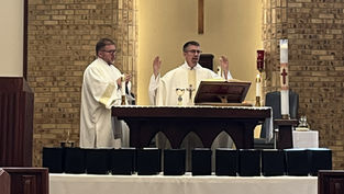 Two clergy in white vestments lead a service at an altar with candles, a cross, and chalice, against a brick wall backdrop, creating a solemn mood.