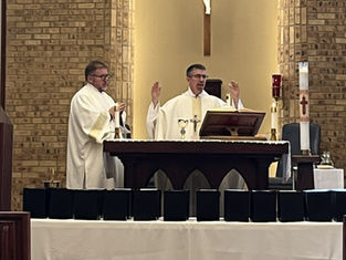 Two clergy in white vestments lead a service at an altar with candles, a cross, and chalice, against a brick wall backdrop, creating a solemn mood.