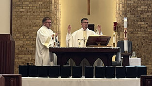 Two clergy in white vestments lead a service at an altar with candles, a cross, and chalice, against a brick wall backdrop, creating a solemn mood.