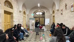 A nun speaks to seated people in a stone room with colorful balloons on patterned floor. An exit sign, posters, and a cross adorn the walls.