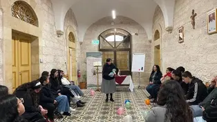 A nun speaks to seated people in a stone room with colorful balloons on patterned floor. An exit sign, posters, and a cross adorn the walls.