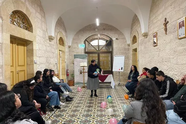 A nun speaks to seated people in a stone room with colorful balloons on patterned floor. An exit sign, posters, and a cross adorn the walls.