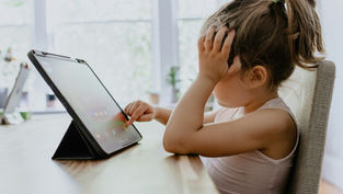 Child in a white top using a tablet, resting head on hand, seated at a wooden table. Bright, blurred background with windows and plants.