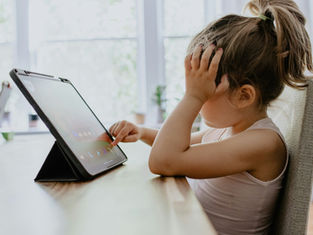 Child in a white top using a tablet, resting head on hand, seated at a wooden table. Bright, blurred background with windows and plants.