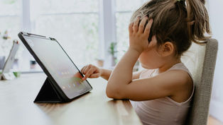 Child in a white top using a tablet, resting head on hand, seated at a wooden table. Bright, blurred background with windows and plants.