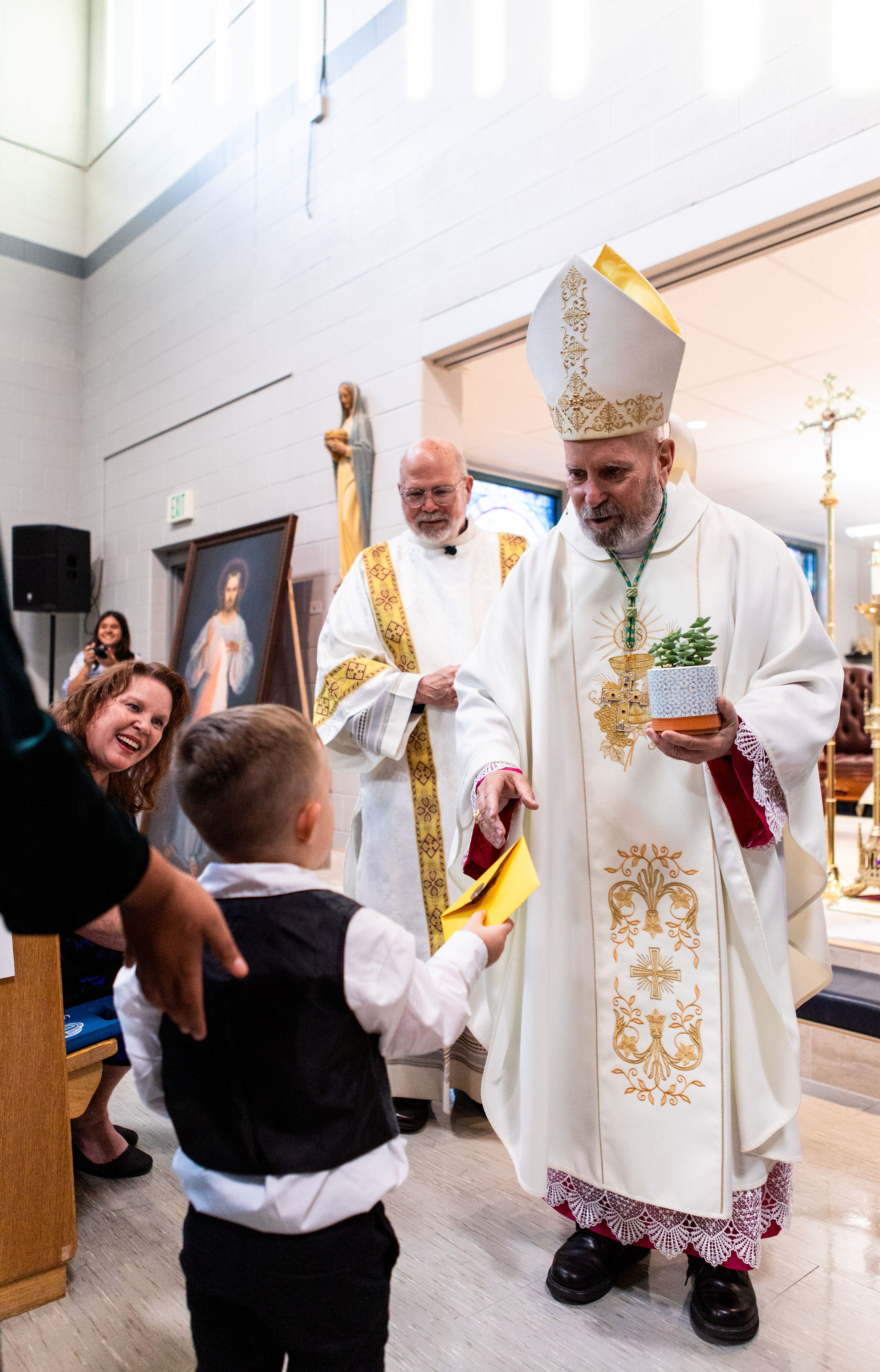Young boy hands yellow envelope to a man in ornate white robe and hat, holding a plant, in a church with statues and paintings. Joyful mood.