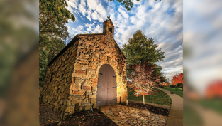 Stone chapel with arched wooden door under a vibrant sky. Surrounded by trees with autumn leaves, creating a serene and peaceful mood.