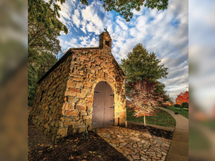 Stone chapel with arched wooden door under a vibrant sky. Surrounded by trees with autumn leaves, creating a serene and peaceful mood.