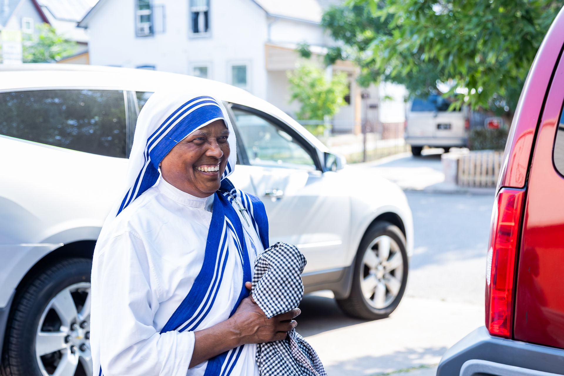 Smiling woman in white and blue robe stands between cars on a sunny street, holding a checkered cloth. Background features trees and houses.