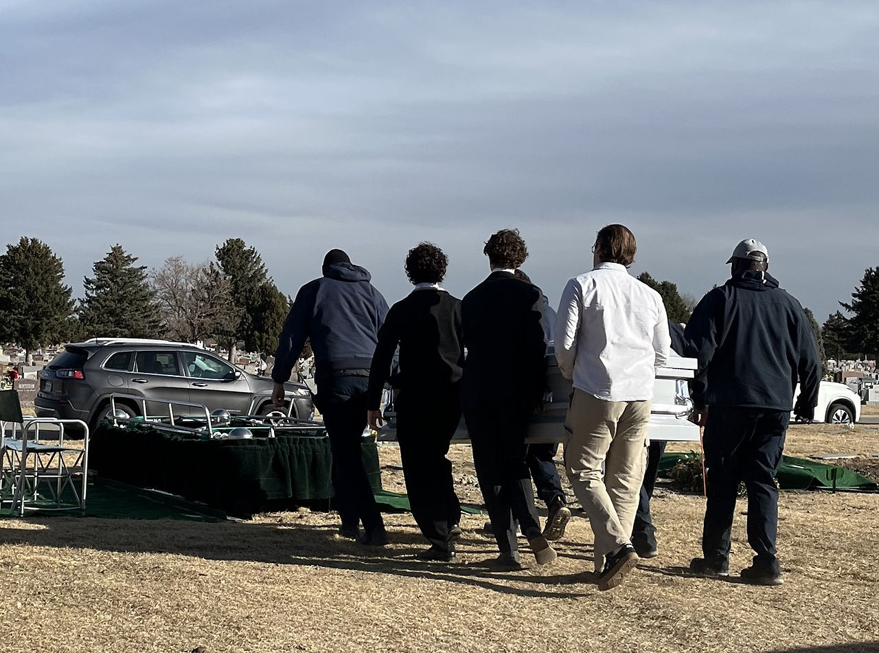 Six people in dark clothing carry a casket across a cemetery with trees and cars in the background, conveying a somber mood.