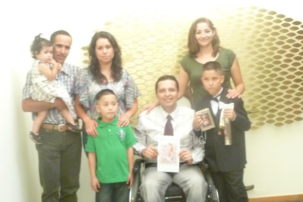 A family of seven poses smiling; a man in a wheelchair holds a photo. One boy in a suit holds a frame and scroll. Honeycomb wall pattern behind.
