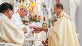 Two clergymen in ceremonial robes perform a ritual. Ornate church interior with flowers in the background. Solemn mood.