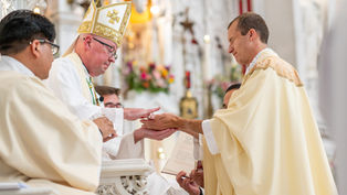 Two clergymen in ceremonial robes perform a ritual. Ornate church interior with flowers in the background. Solemn mood.