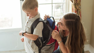 Mother helps young boy put on a backpack by a bright window, preparing him for school.