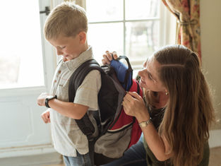 Mother helps young boy put on a backpack by a bright window, preparing him for school.