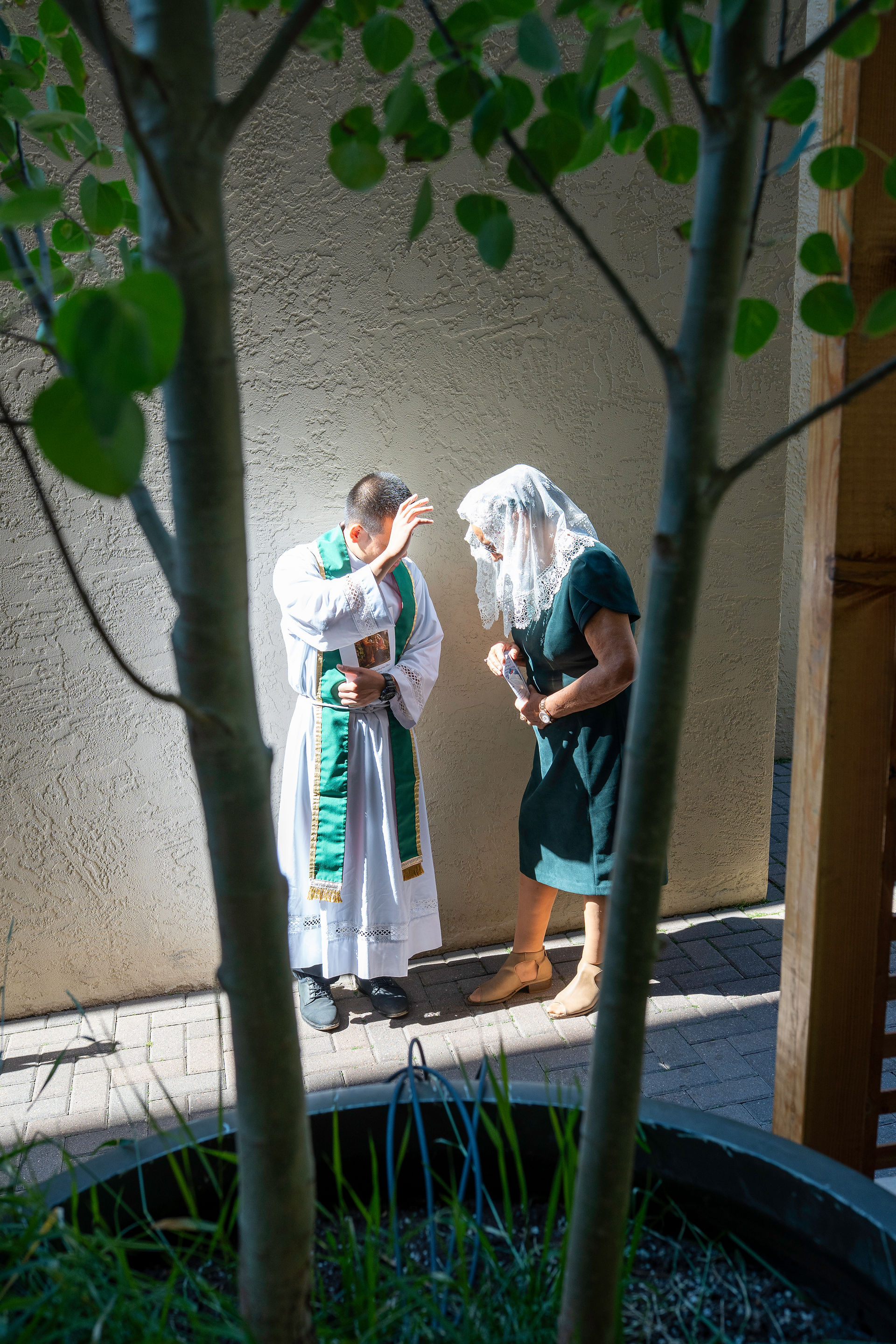 Two individuals stand outdoors near a wall, partially obscured by tree branches. One wears religious vestments with green and gold accents and gestures with the right hand; the other wears a dark dress and a white lace veil.