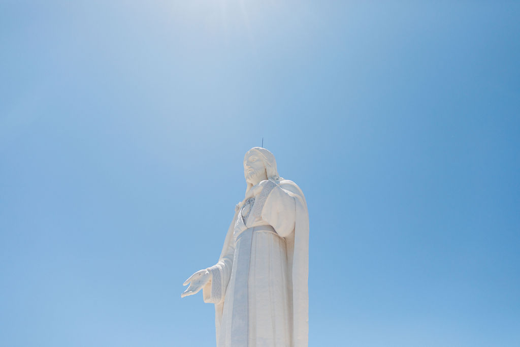 White statue of Jesus with outstretched hand against a bright blue sky and sun glare, serene and uplifting