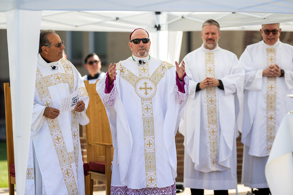 Four priests in white-and-gold vestments stand under a canopy; the center priest speaks with raised hands at an outdoor ceremony.