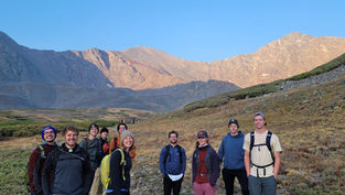 Smiling group of hikers with backpacks on a mountain trail under sunny blue sky, surrounded by rocky peaks and tundra.