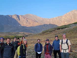 Smiling group of hikers with backpacks on a mountain trail under sunny blue sky, surrounded by rocky peaks and tundra.