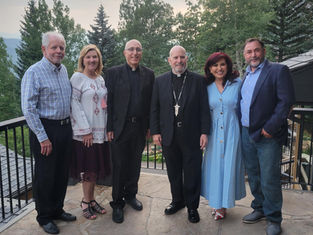 Group of six people smiling on a stone patio, wearing formal and casual attire, with lush green trees and overcast sky in the background.