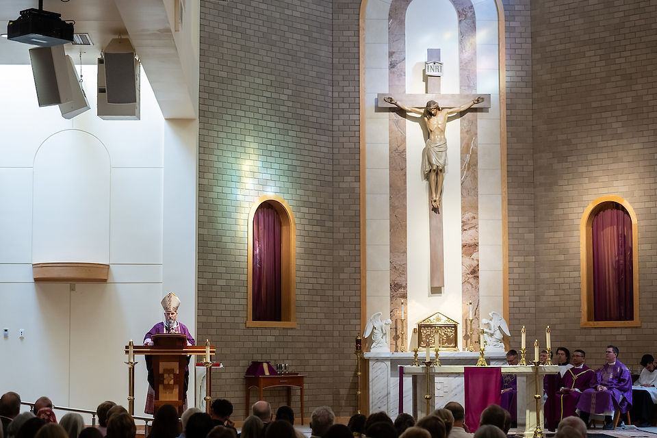 Bishop in purple robes speaks at a pulpit in a church, with a crucifix, altar, and audience. Warm lighting creates a solemn atmosphere.