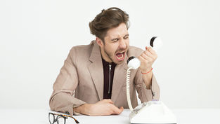A man in a beige suit angrily yells into a white rotary phone. Glasses are on the table in a neutral white setting. Emotion: frustration.