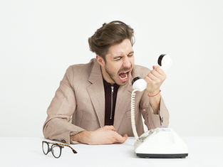 A man in a beige suit angrily yells into a white rotary phone. Glasses are on the table in a neutral white setting. Emotion: frustration.