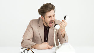 A man in a beige suit angrily yells into a white rotary phone. Glasses are on the table in a neutral white setting. Emotion: frustration.