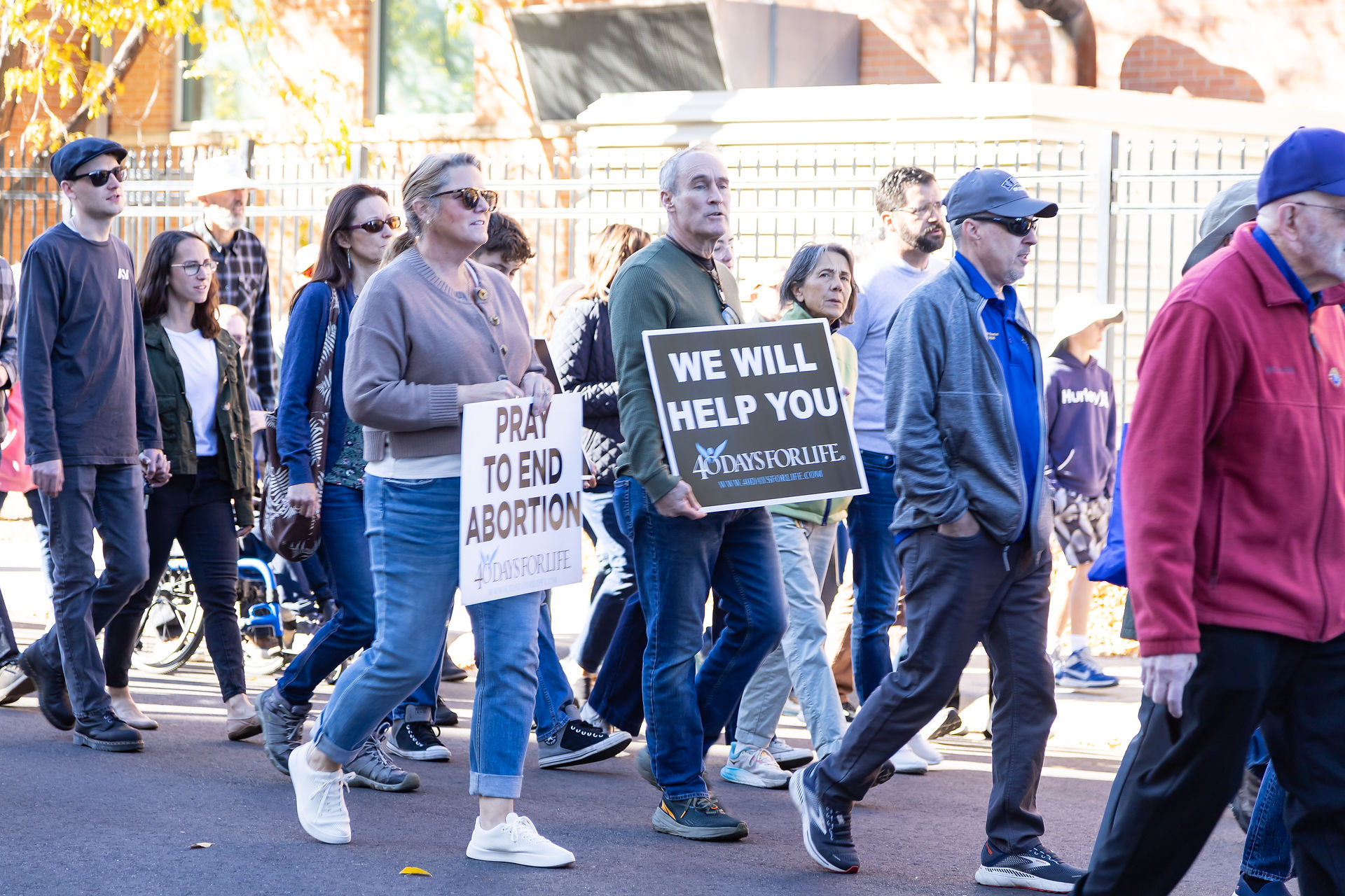 People march holding pro-life signs reading "Pray to End Abortion" and "We Will Help You," against a sunny urban backdrop.