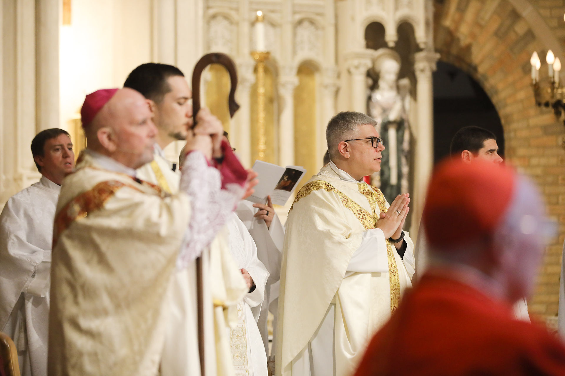 Members of the clergy pray during a religious ceremony. The background features an ornate ivory and gold altar. The mood is celebratory.