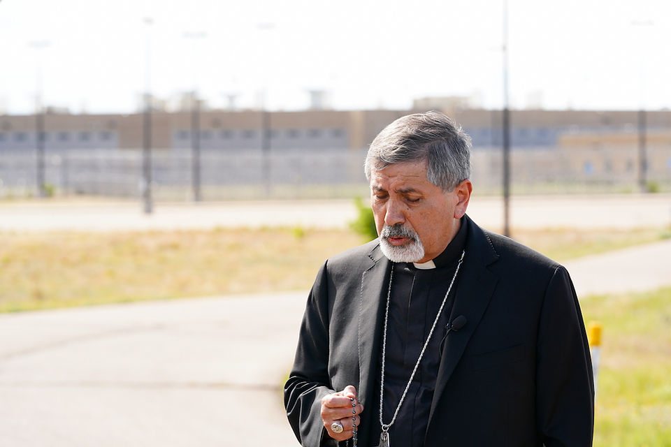 Catholic priest in black clerical clothes holds a rosary outdoors, looking down beside a road and fenced buildings.
