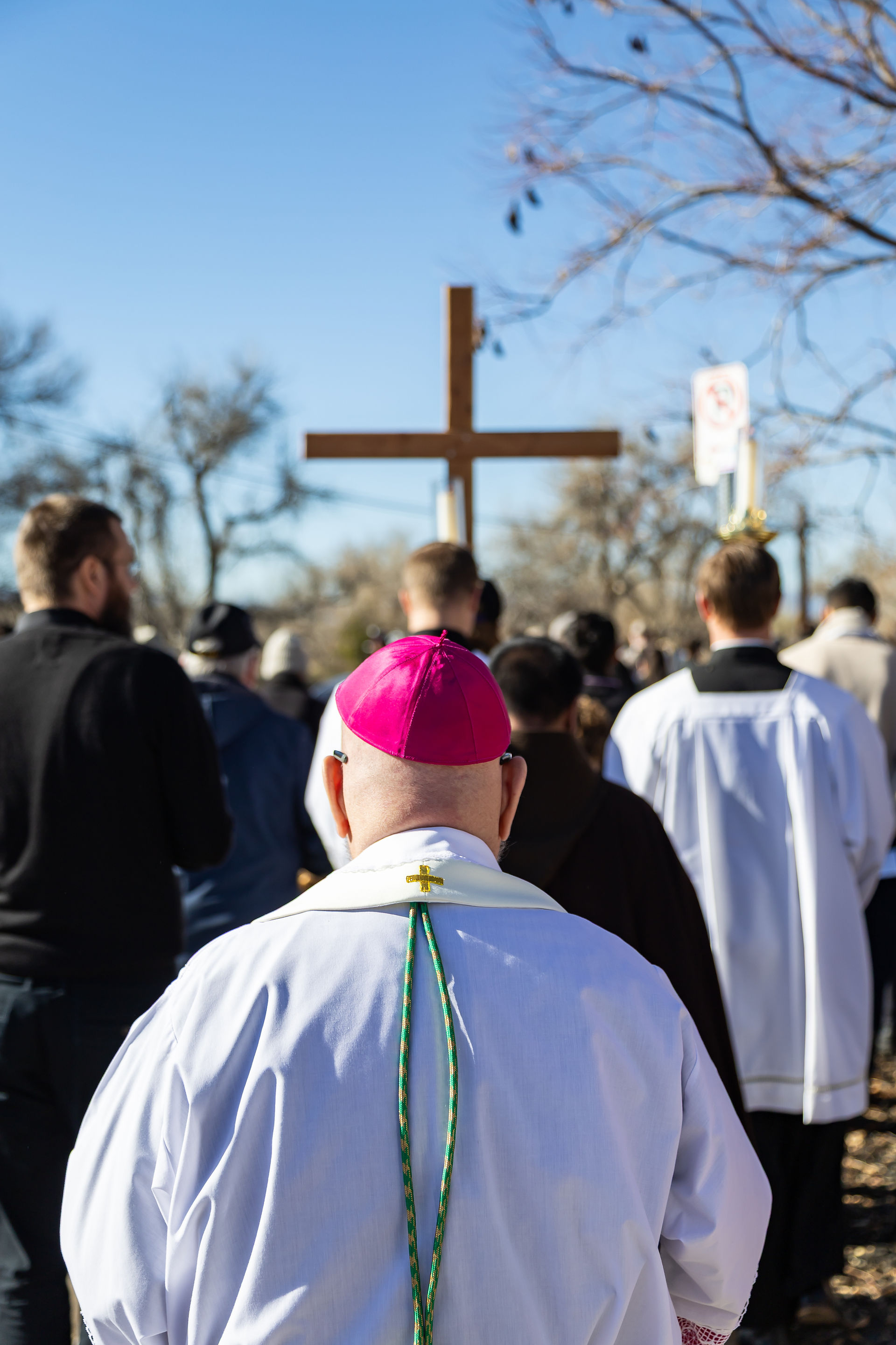 A group in religious attire walks toward a large wooden cross outdoors. A man in a pink cap leads. Bare trees and a clear sky set the scene.