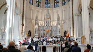 People gathered in a church near an ornate altar with stained glass windows. A priest addresses the congregation in a ceremonial setting.