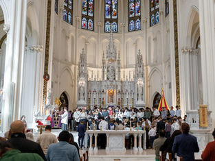 People gathered in a church near an ornate altar with stained glass windows. A priest addresses the congregation in a ceremonial setting.