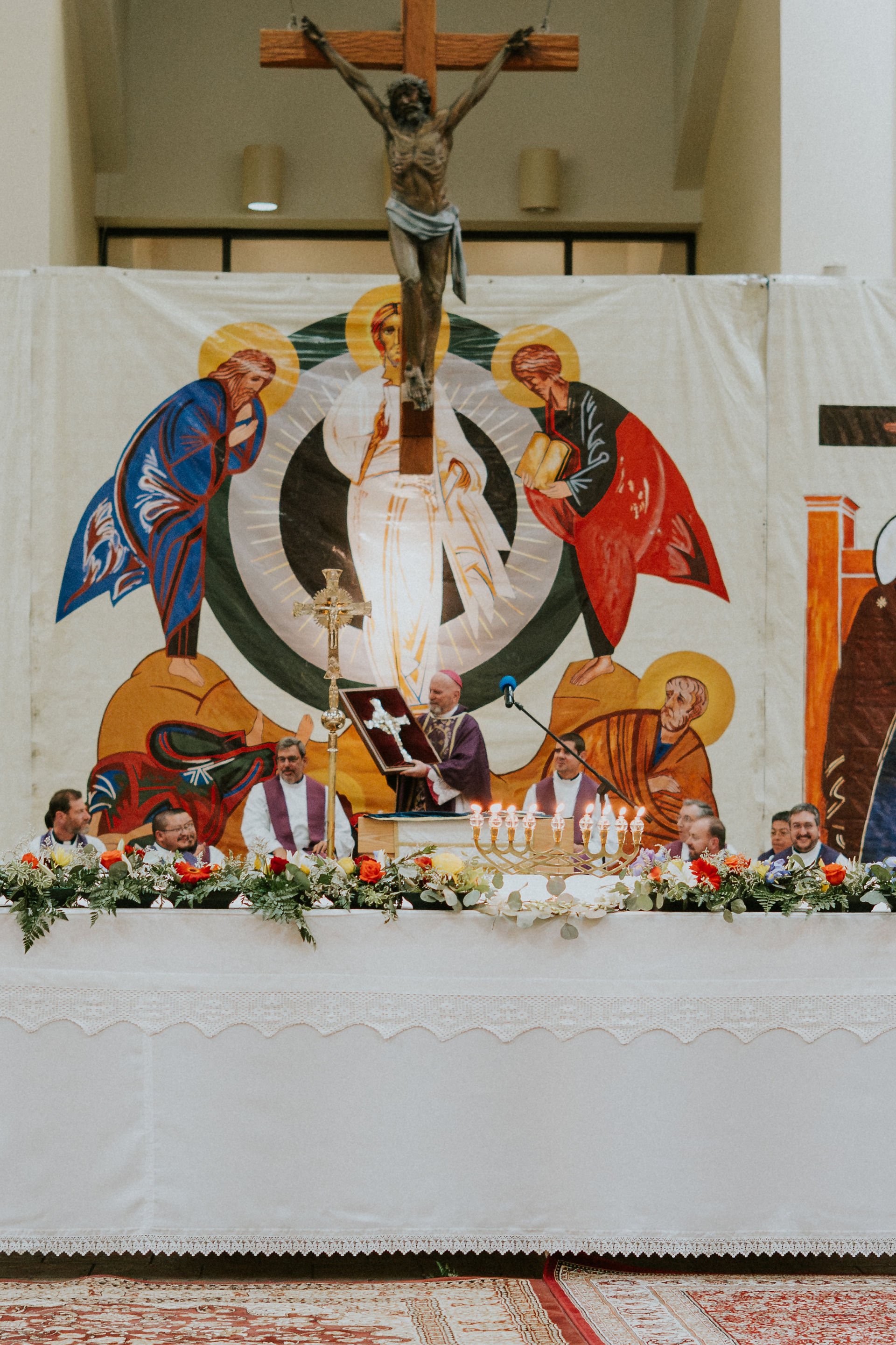 Clergy gather behind an altar with flowers and candles, holding a cross plaque. A tapestry with religious figures and crucifix is in the background.