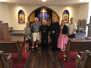 Group of six people standing inside a church with wooden pews and religious icons. Two clergy members in black, others in casual clothing.