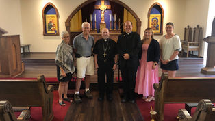Group of six people standing inside a church with wooden pews and religious icons. Two clergy members in black, others in casual clothing.
