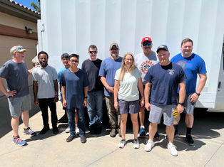 Group of 10 people pose smiling outdoors beside a white moving truck, with a sunny driveway and house in the background.