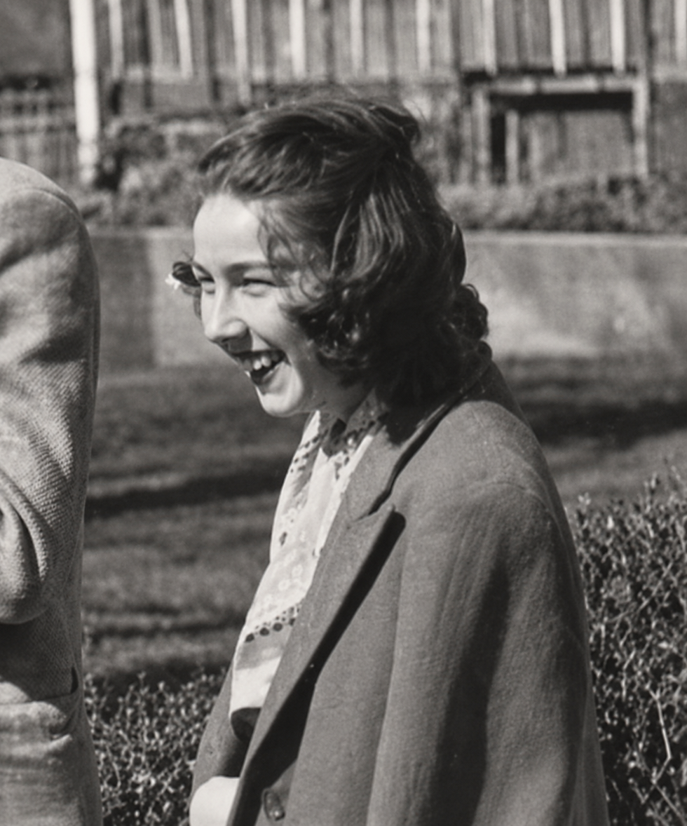 Black-and-white photo of a young woman laughing outdoors, with a building and hedges behind her and a partial person at left.
