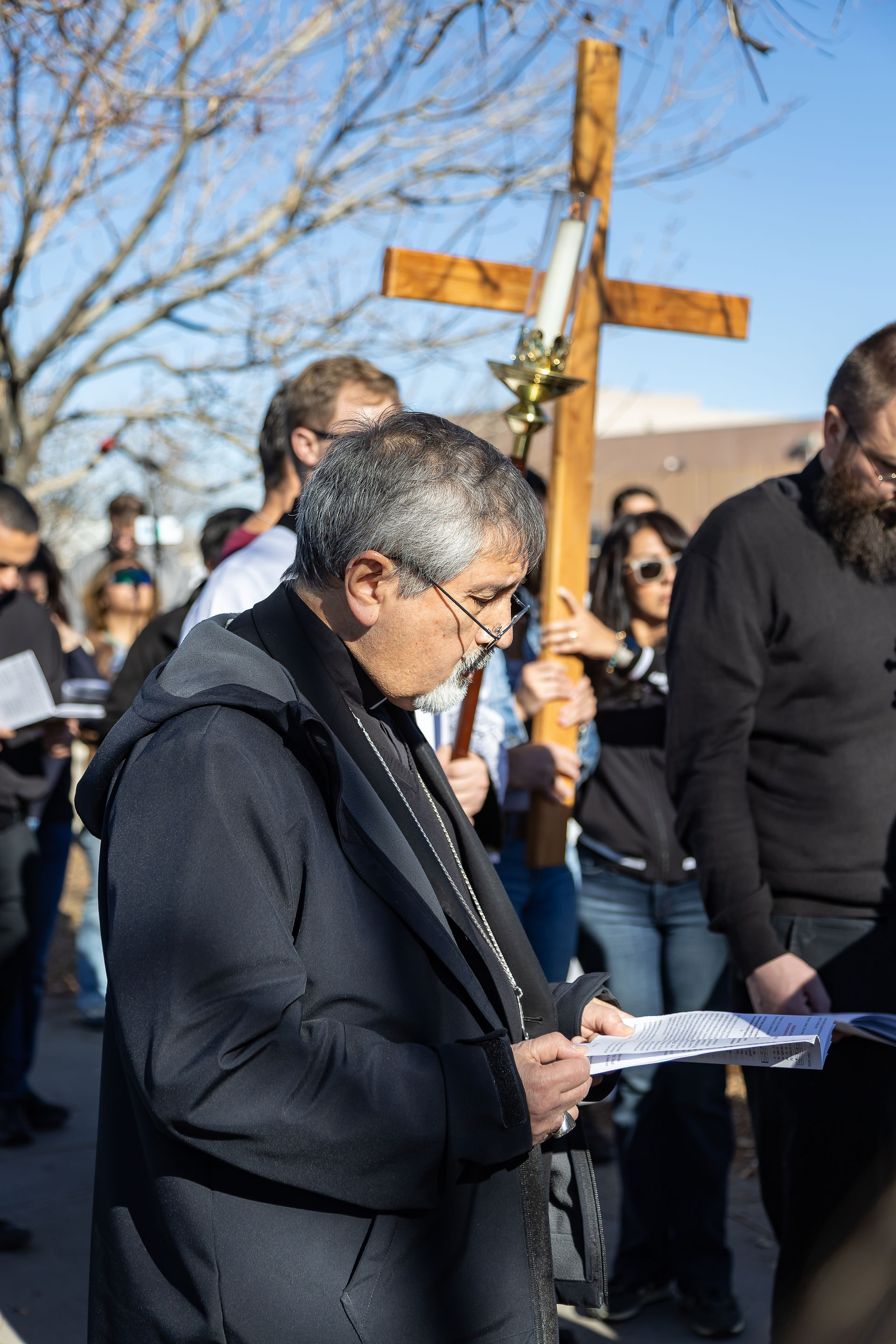 People hold a cross and papers during an outdoor gathering on a sunny day. One person looks down in focus. Background of bare trees.