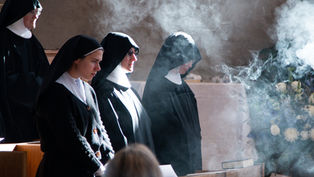 Nuns in black habits stand solemnly in a church amidst incense smoke, with sunlight highlighting their expressions, creating a serene atmosphere.