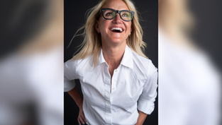 Smiling woman with glasses and blonde hair in a white shirt against a dark background, looking up with joy.