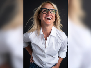 Smiling woman with glasses and blonde hair in a white shirt against a dark background, looking up with joy.