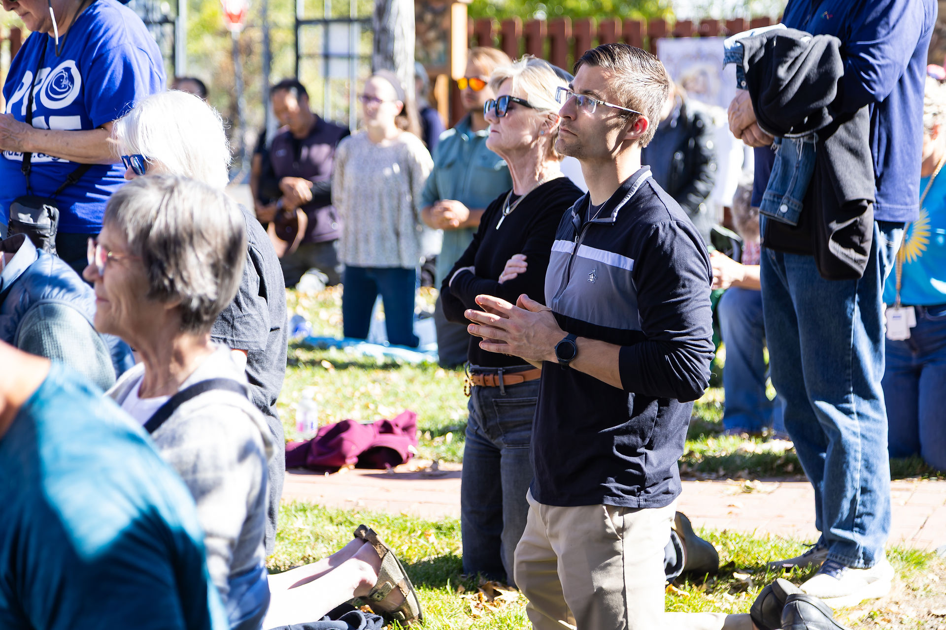 People are kneeling and standing in a park, praying. A man with glasses is in focus, wearing a black top. Sunlight illuminates the scene.