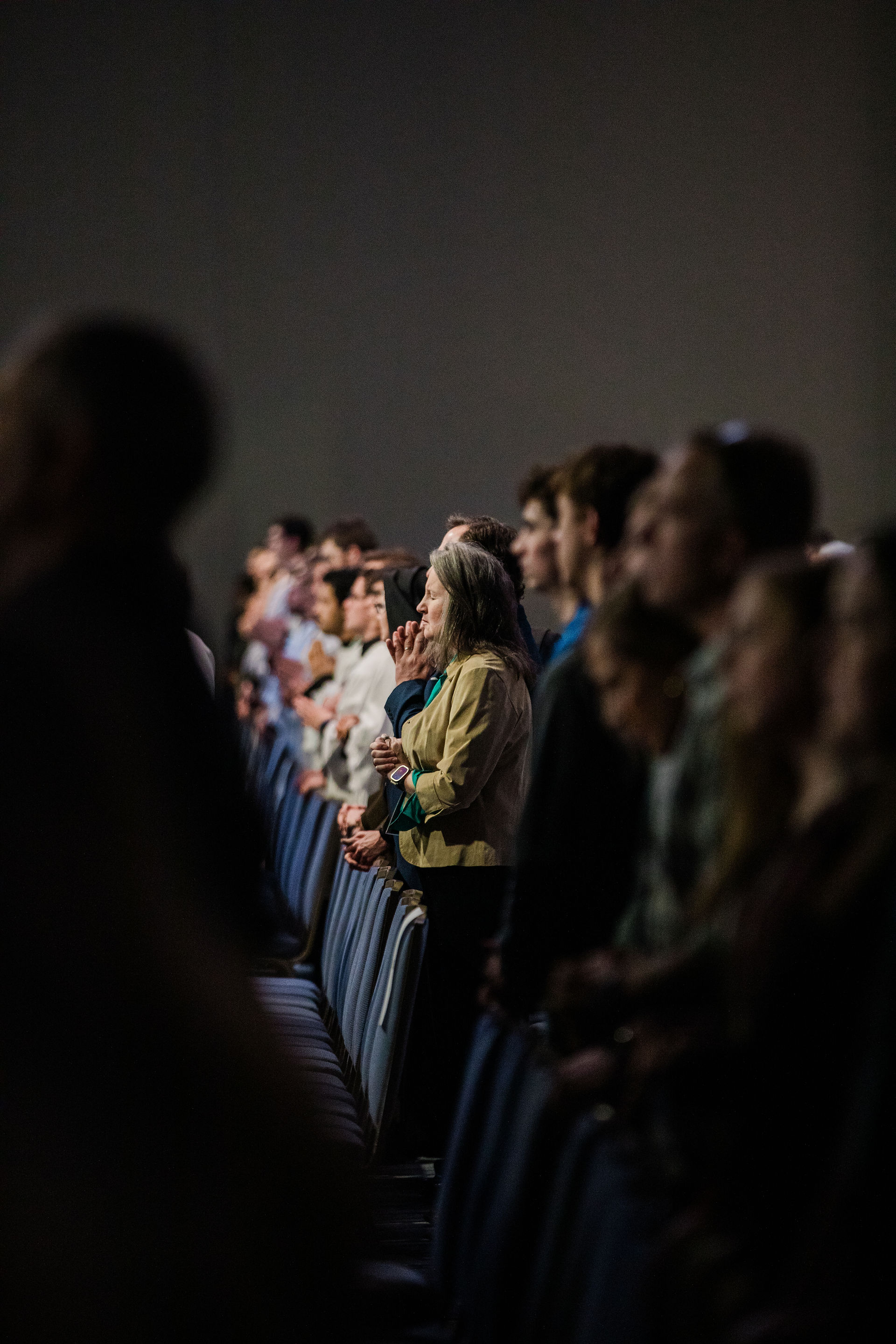 Audience members stand closely together in a dimly lit hall, with one woman in focus holding her hands in prayer while others stand attentively around her.