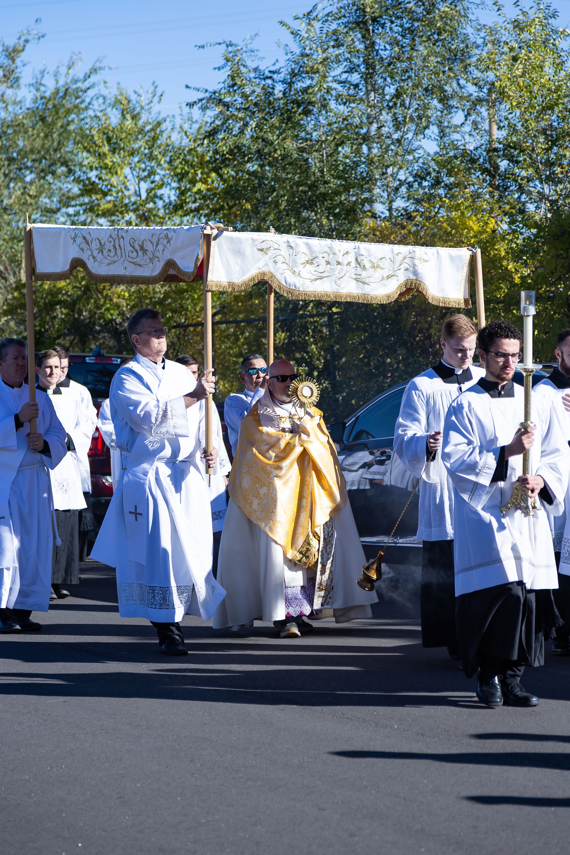 Procession of men in white robes, one holding gold object, under embroidered canopy outdoors. Trees in background, solemn atmosphere.